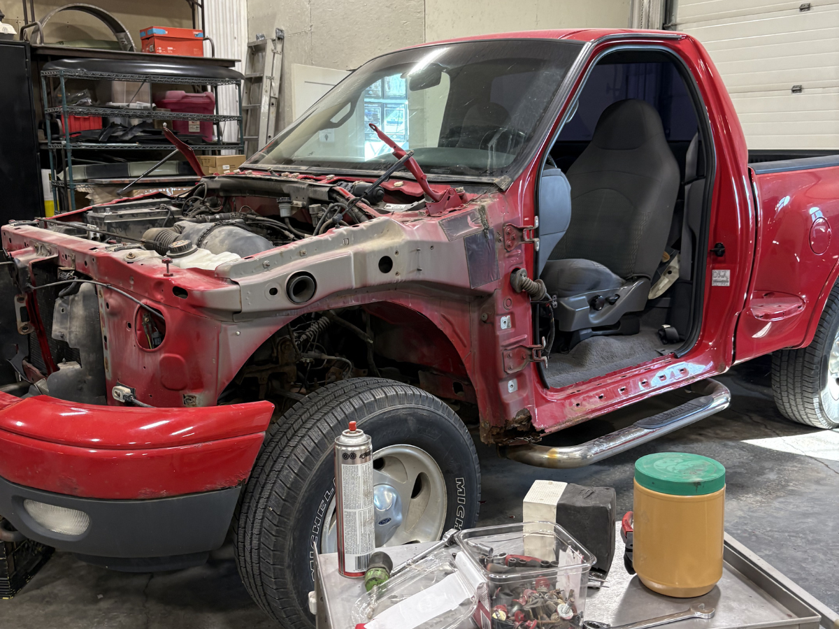 Red pickup truck in a garage with its front end disassembled, showing exposed engine and interior.