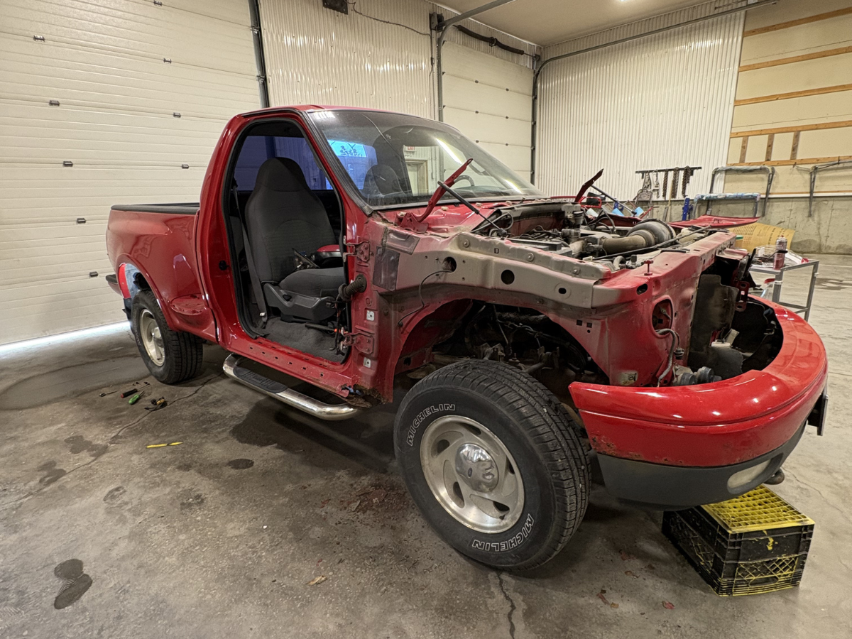 Red pickup truck with missing front end and open door in a garage, undergoing repair