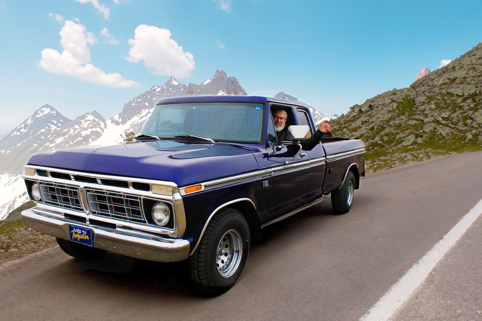 Classic dark blue pickup truck driving on a mountain road with a person leaning out the window.