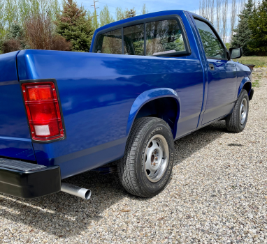 A blue truck with a red tail light is parked on gravel.