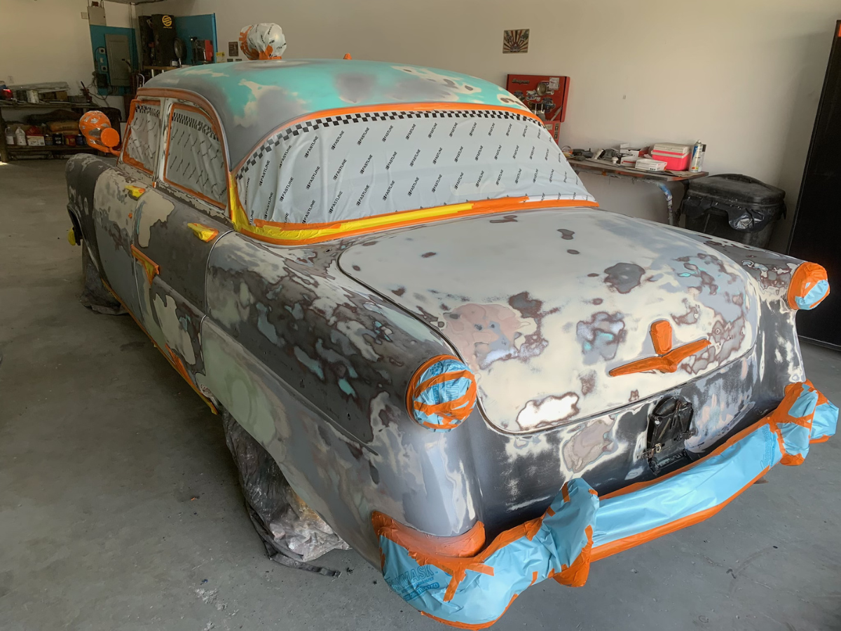 Primered classic car in a garage, covered with orange masking tape and bodywork patches.