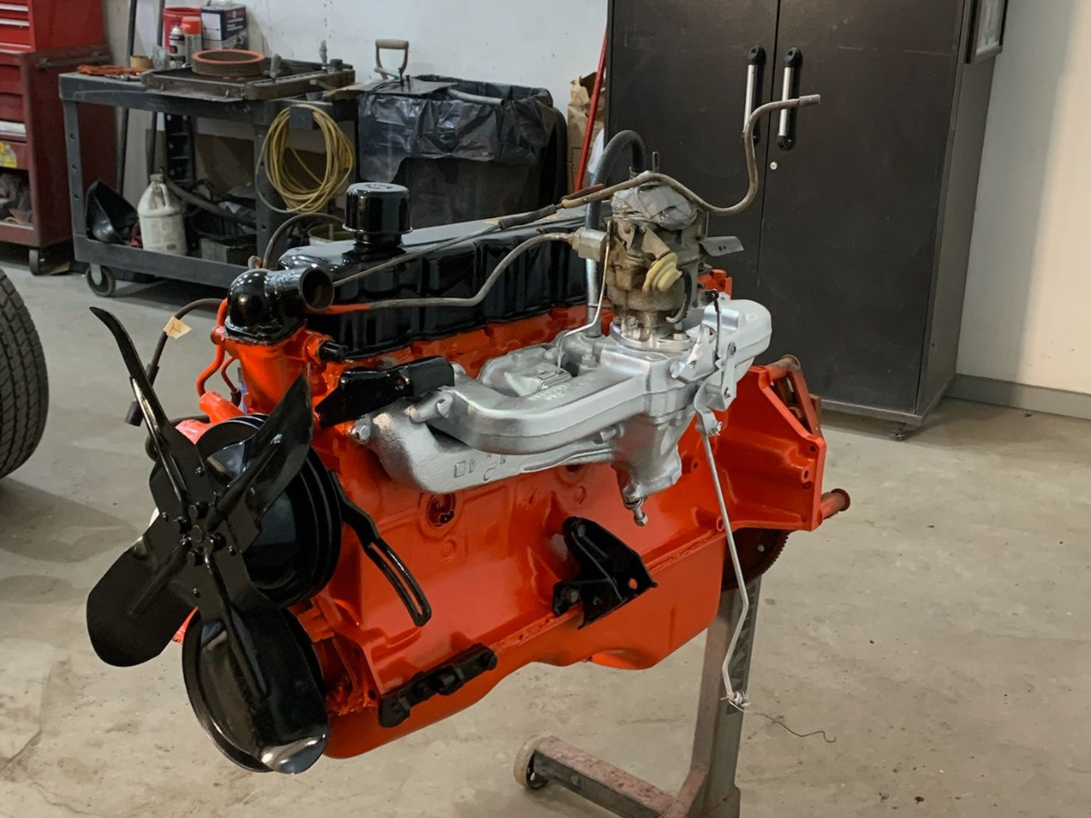 Orange car engine on a stand in a workshop, with hoses and parts visible in the background.