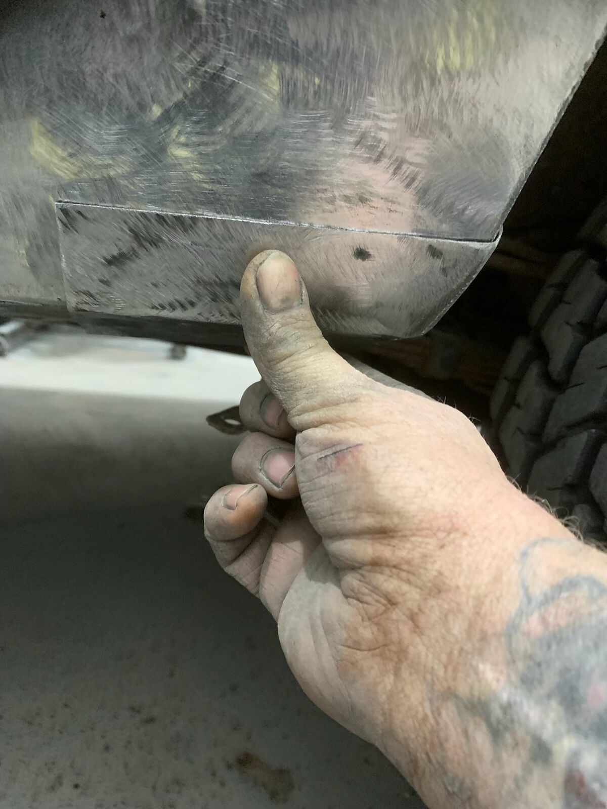A hand points to a metal panel seam under a vehicle, showing a small screw hole and worn surface.
