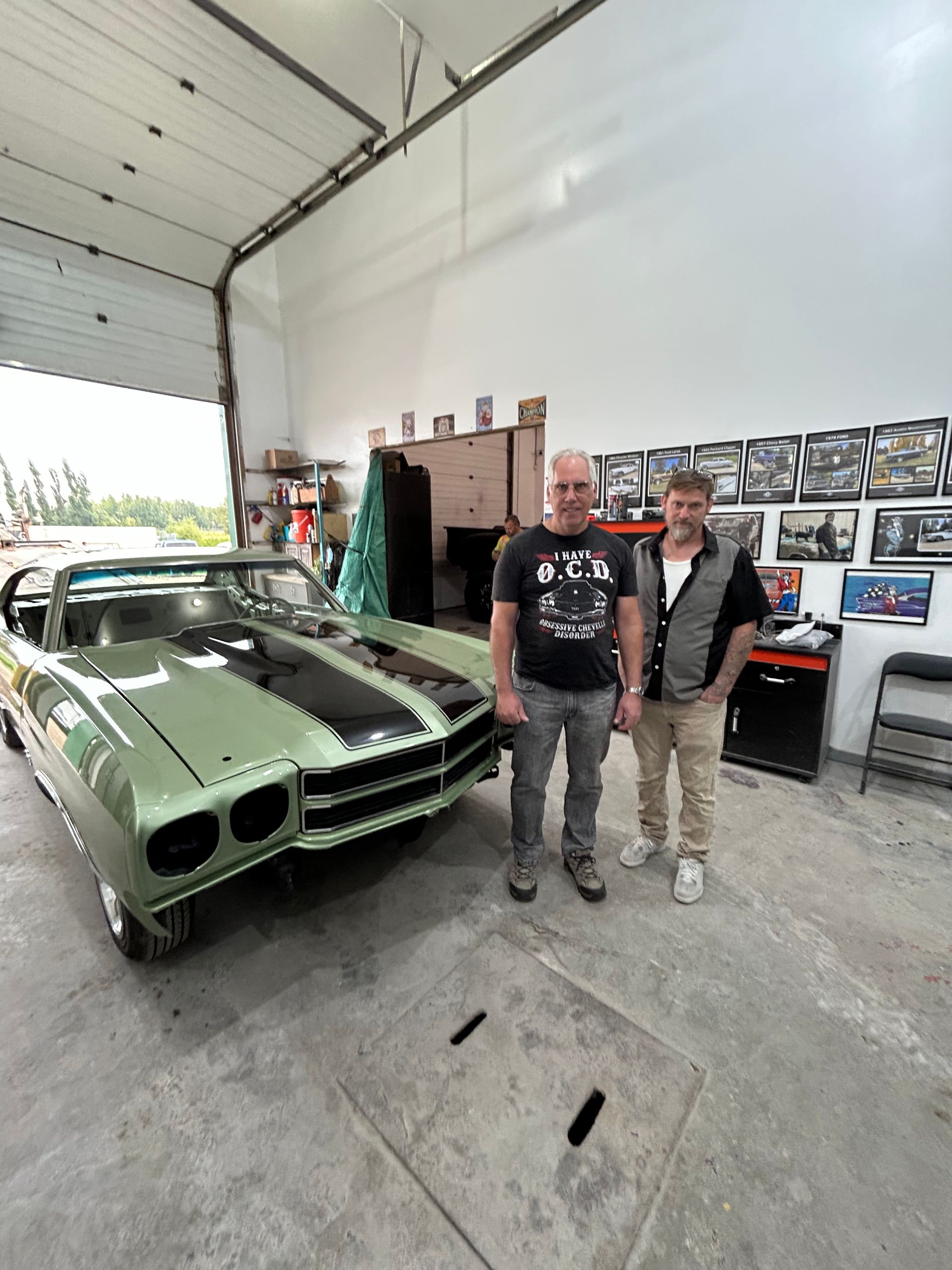 Two men are standing in front of a green car in a garage.