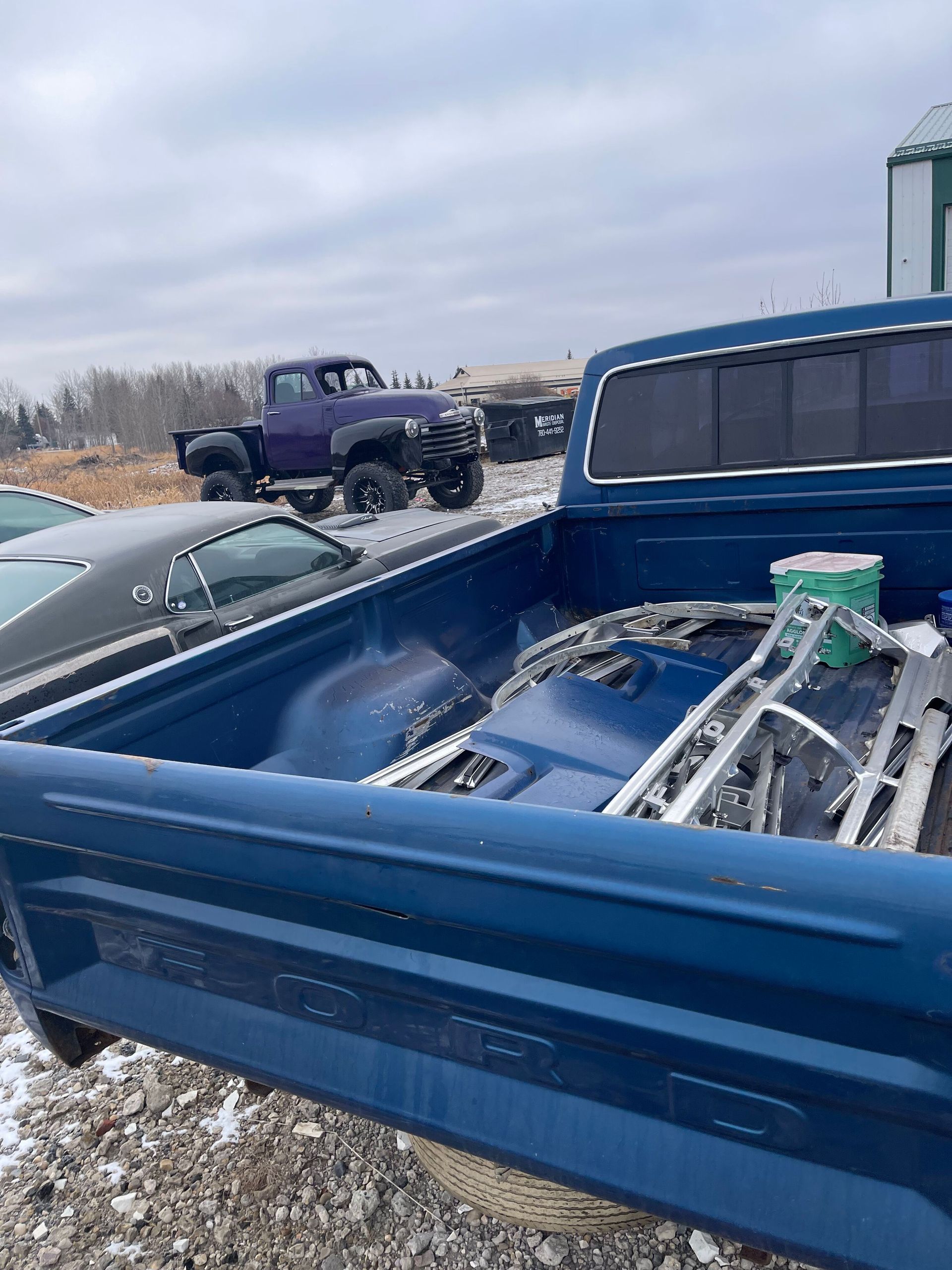 Blue pickup truck bed filled with metal frames and tools in a gravel lot under a cloudy sky