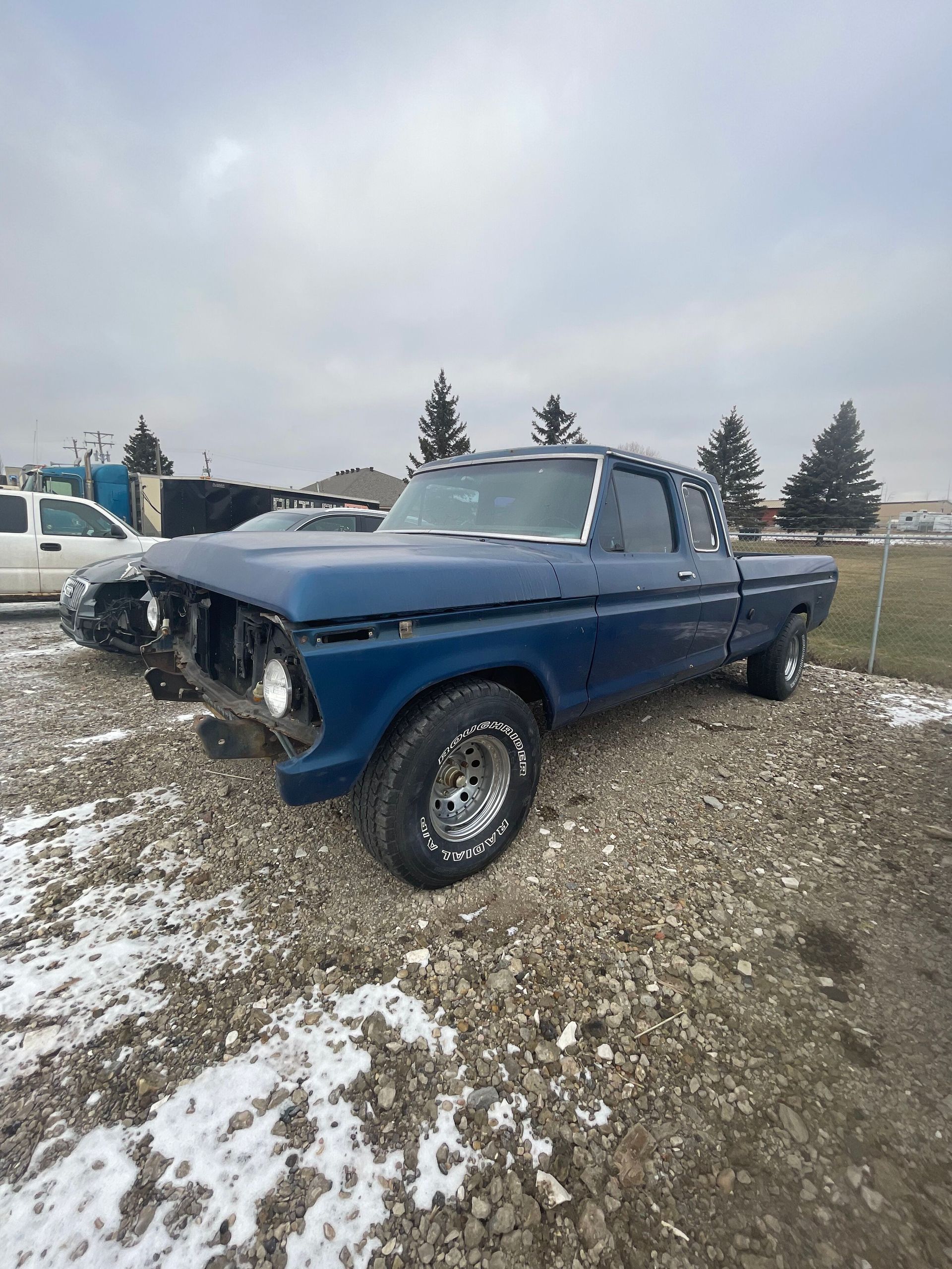 Blue vintage pickup truck parked on gravel with patches of snow under a cloudy sky