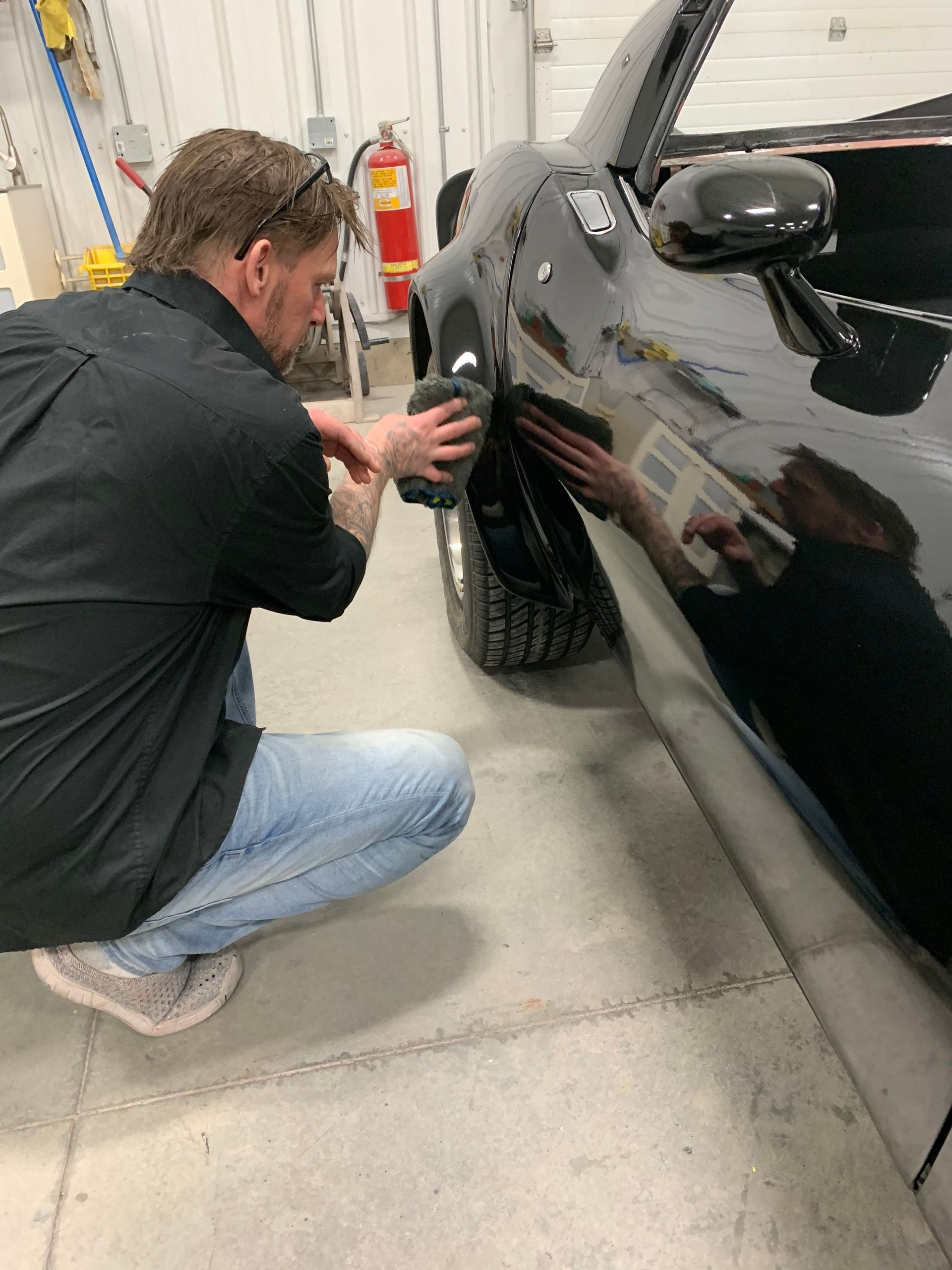 Mechanic kneeling beside a black car, working on the front wheel in a garage.