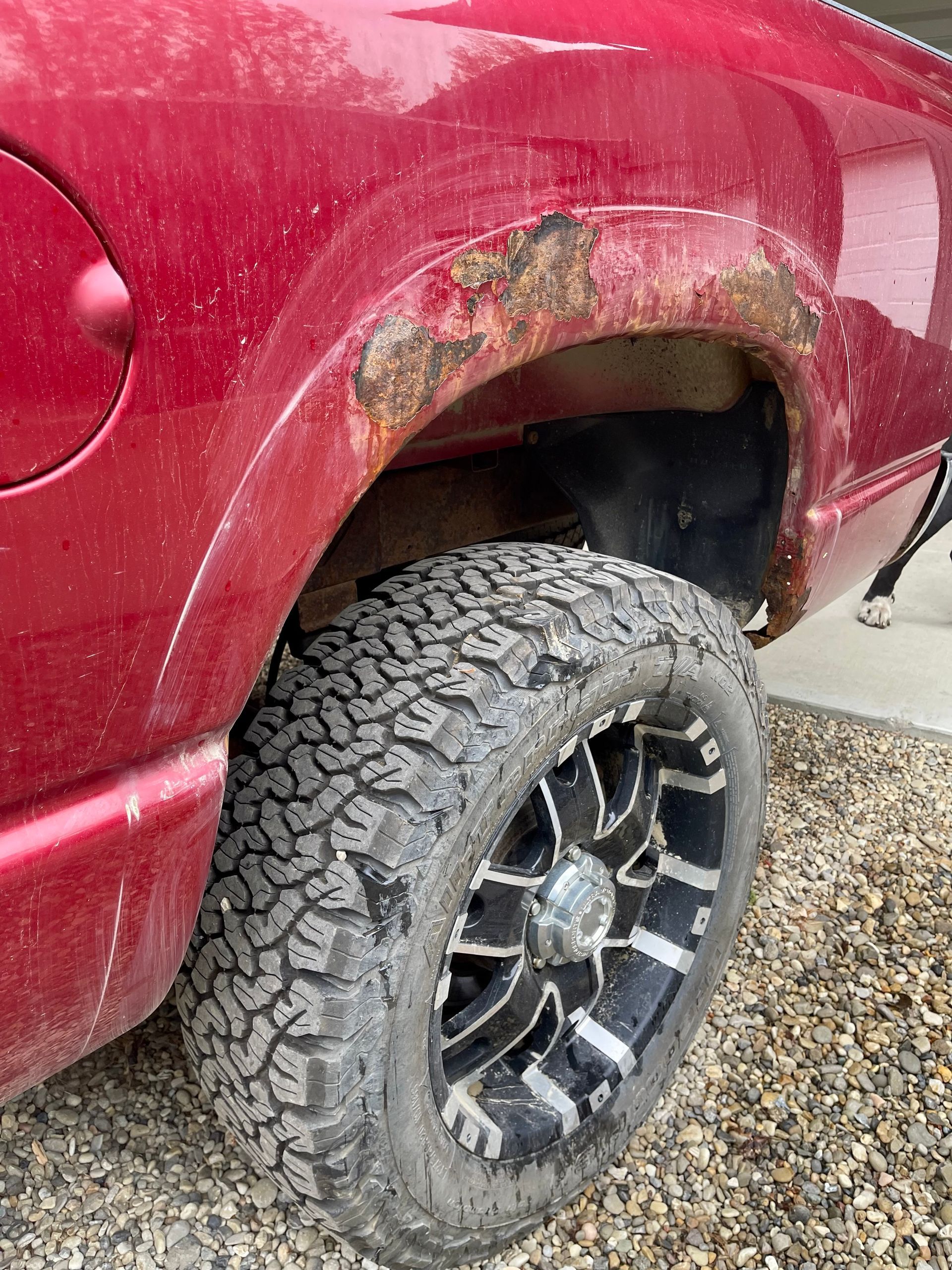 A red truck with rust on the fenders and tires is parked on gravel.