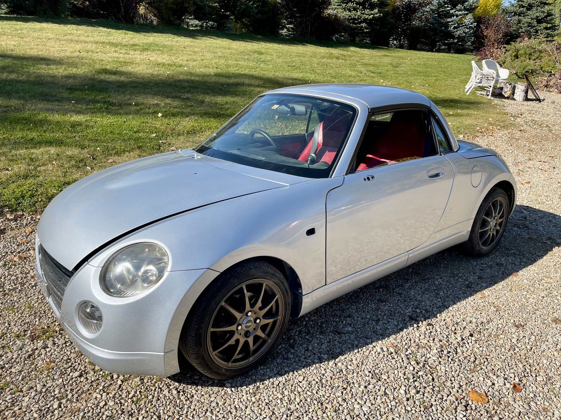 A silver sports car with a red seat is parked in a gravel driveway.