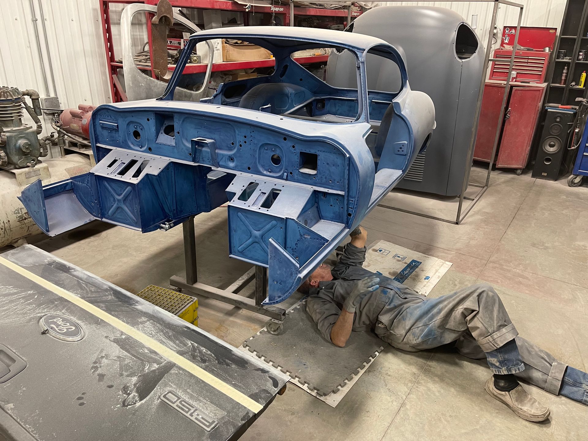 A man is laying on the floor next to a blue car in a garage.