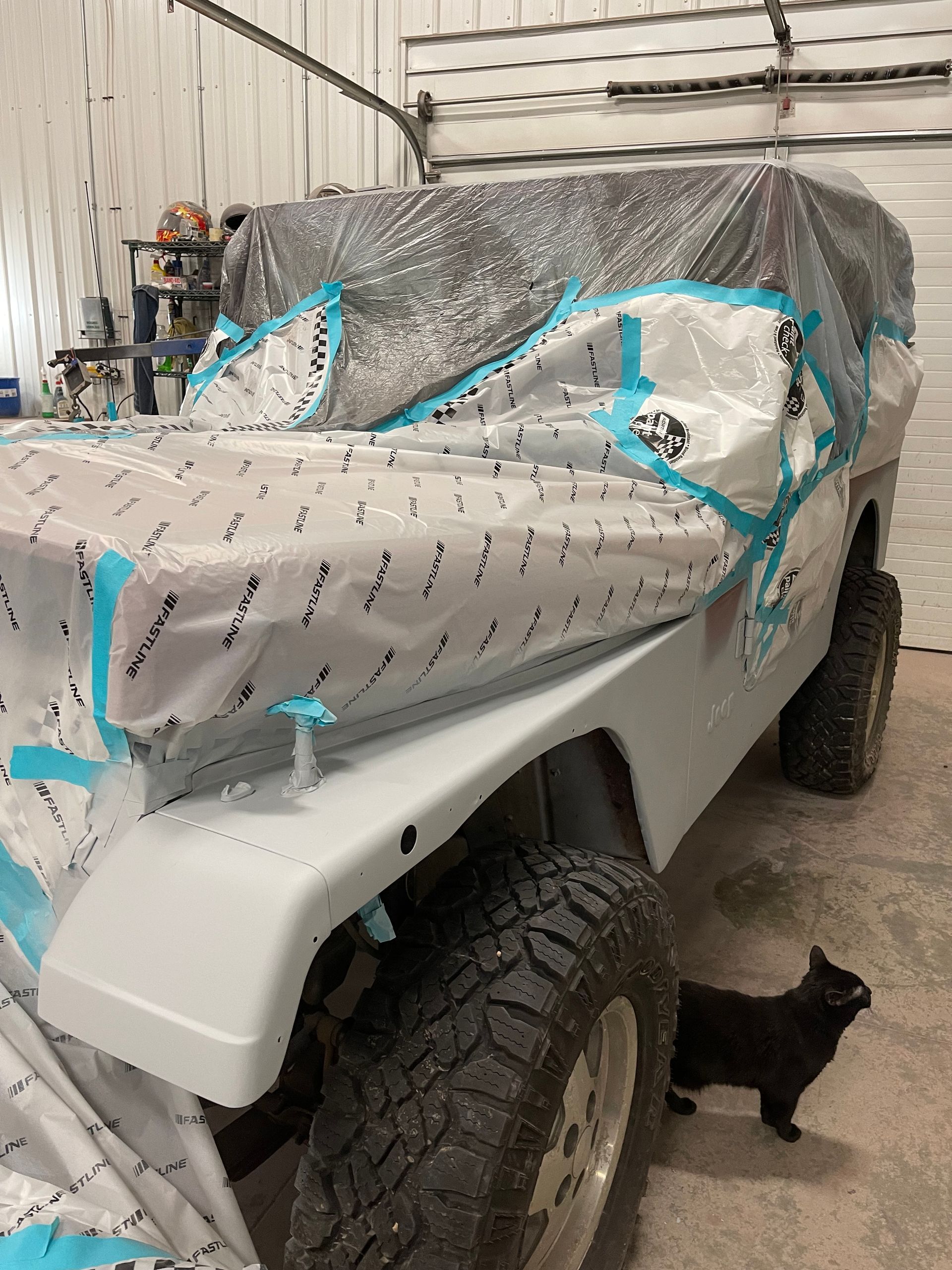 A black cat is standing next to a jeep that is covered in plastic wrap.