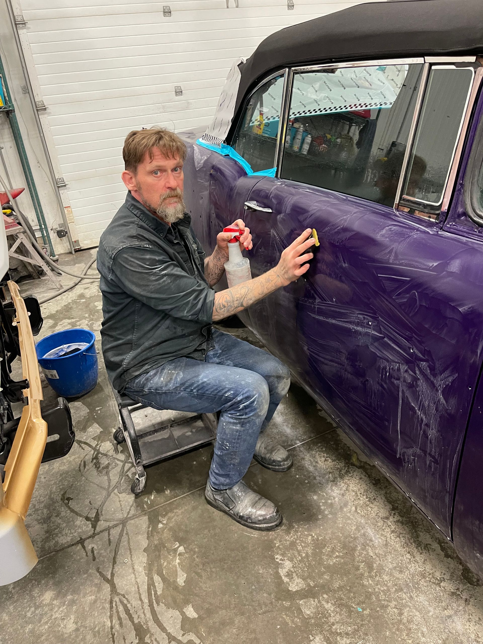 A man is sitting on a stool cleaning a purple car.