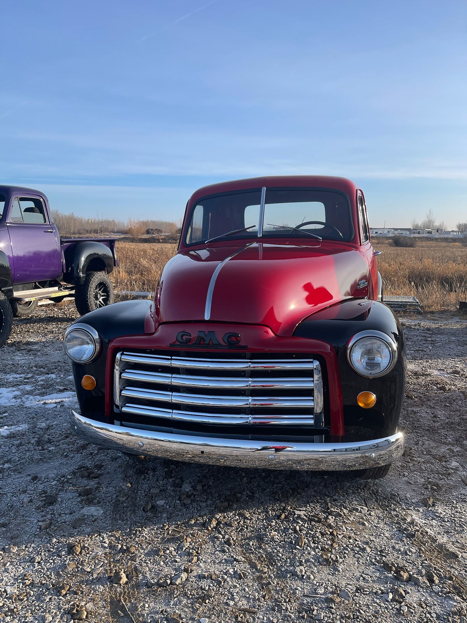 A red truck is parked in a gravel lot next to a purple truck.