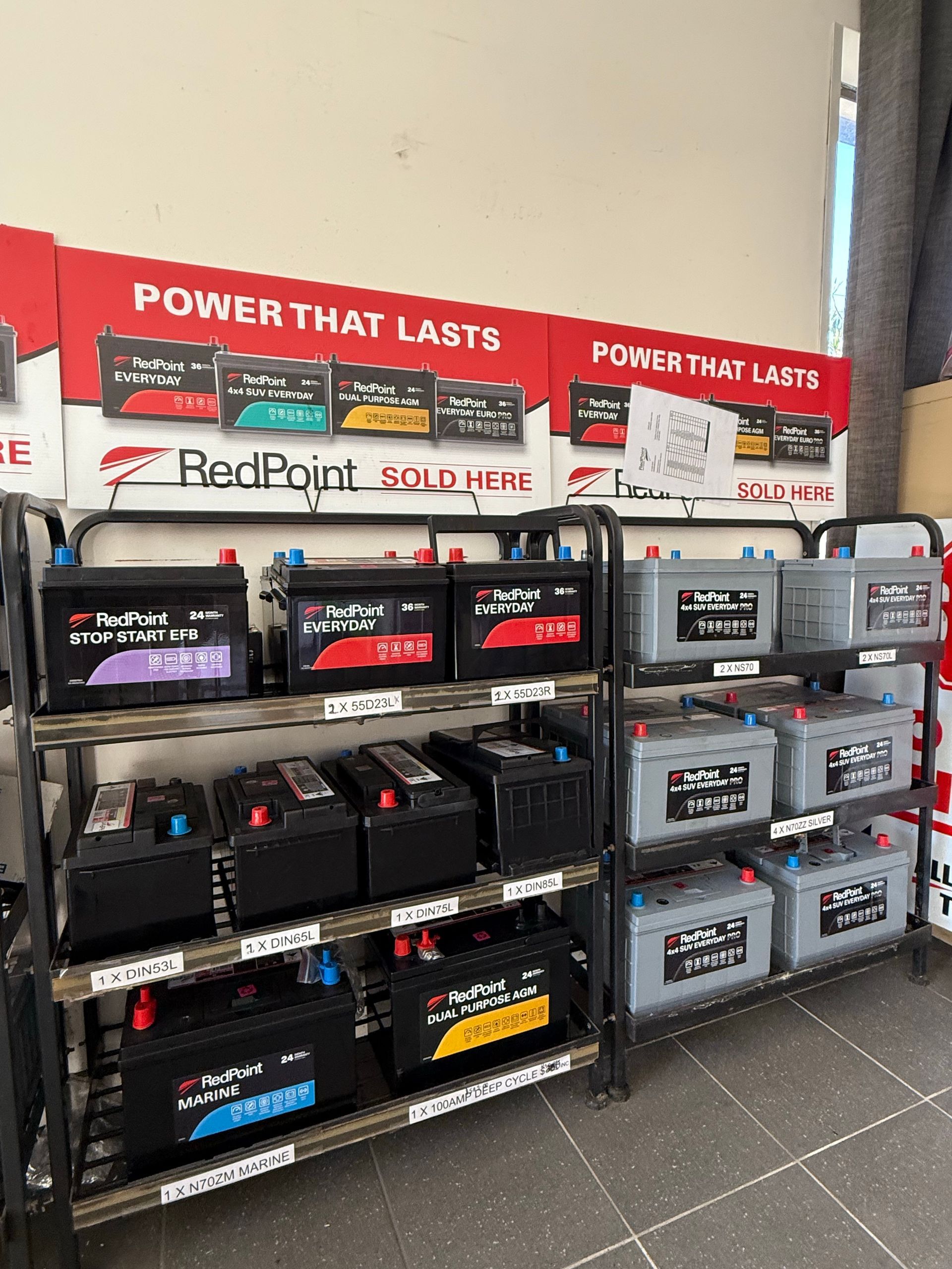 Display of RedPoint car batteries on metal shelves in a store aisle with red “POWER THAT LASTS” signs.