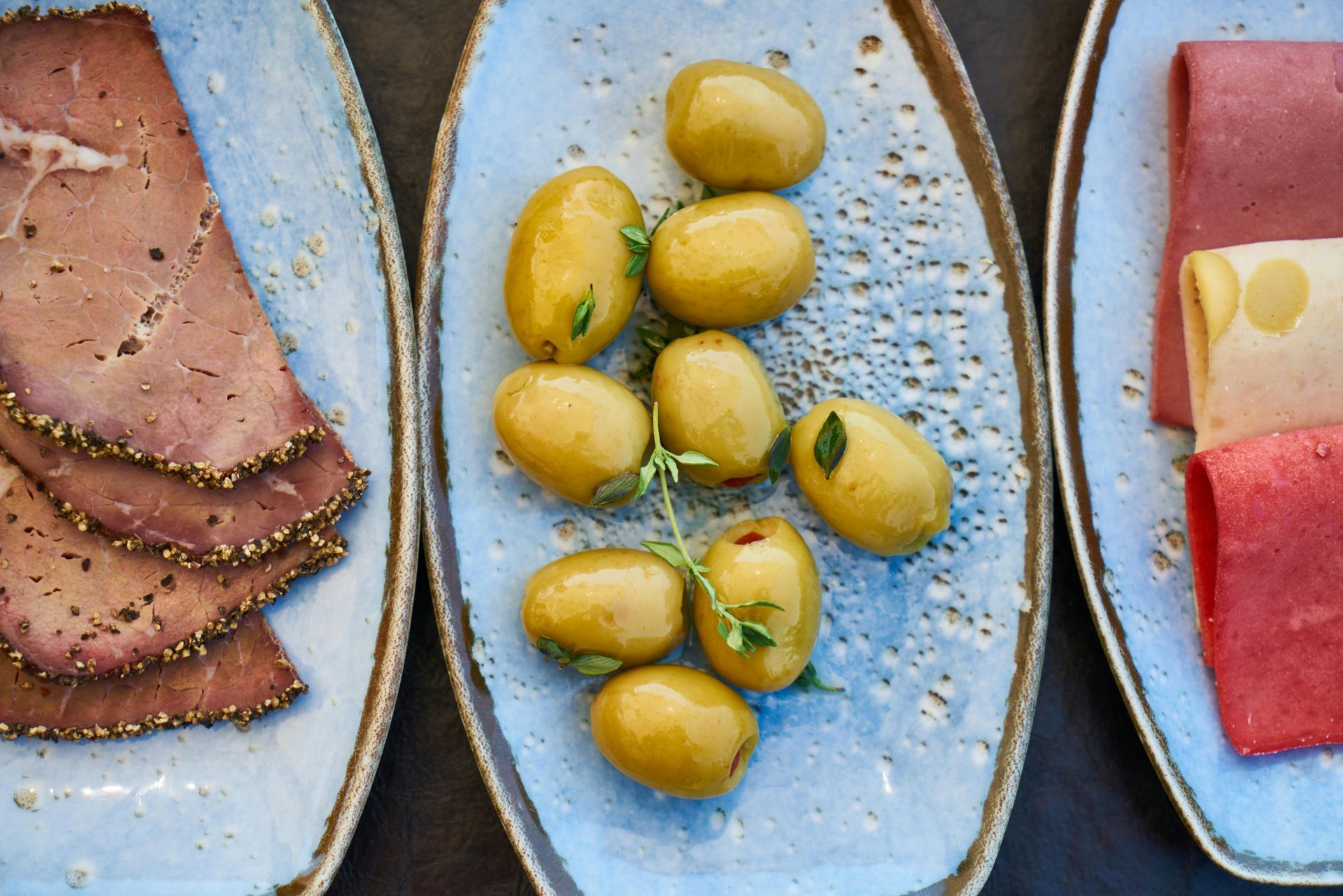 Tallrikar med mat: skivat kött, gröna oliver med örter och mer kött, arrangerade på en mörk yta.