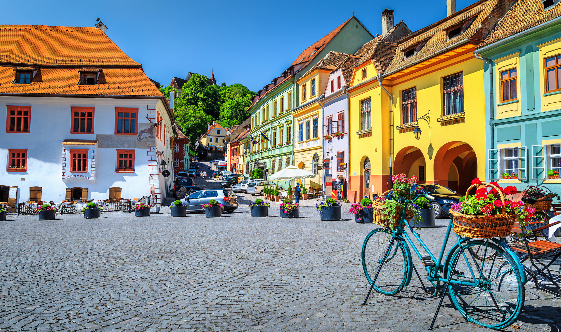 Kullerstenstorg i en europeisk stad med färgglada byggnader. En blå cykel med blomkorgar står i förgrunden.