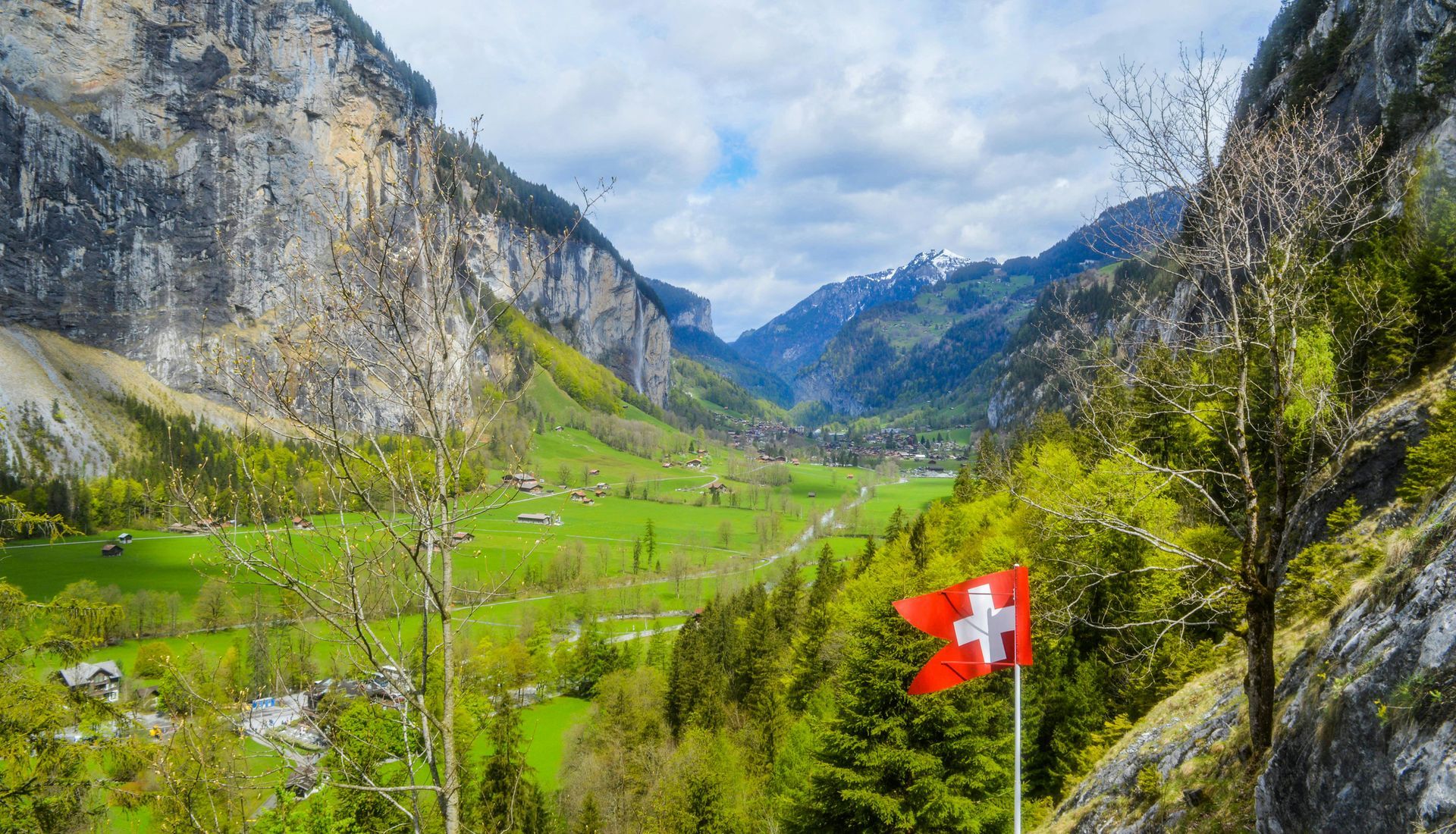 Schweizisk flagga i förgrunden, med utsikt över en frodig dal mellan höga berg.