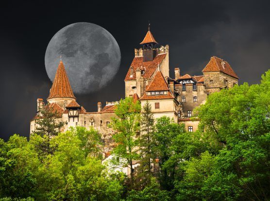 Bran slott, Transsylvanien, med en fullmåne bakom sig. Slottet ligger på en kulle, omgivet av gröna träd.
