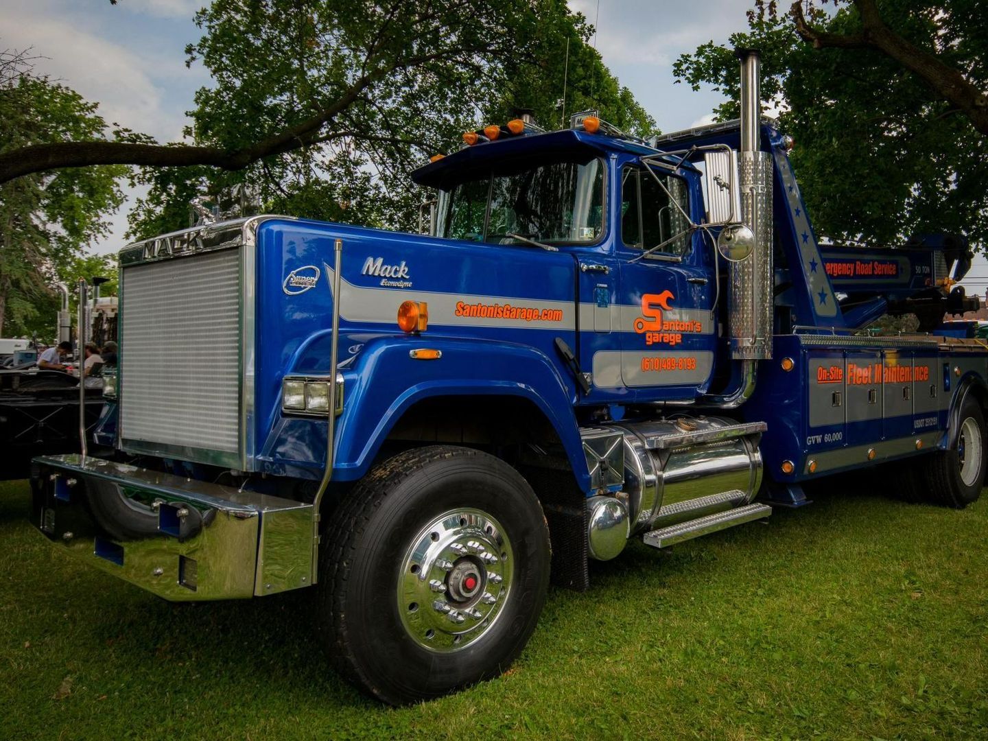 Blue Mack tow truck parked on green grass, chrome accents, under tree.