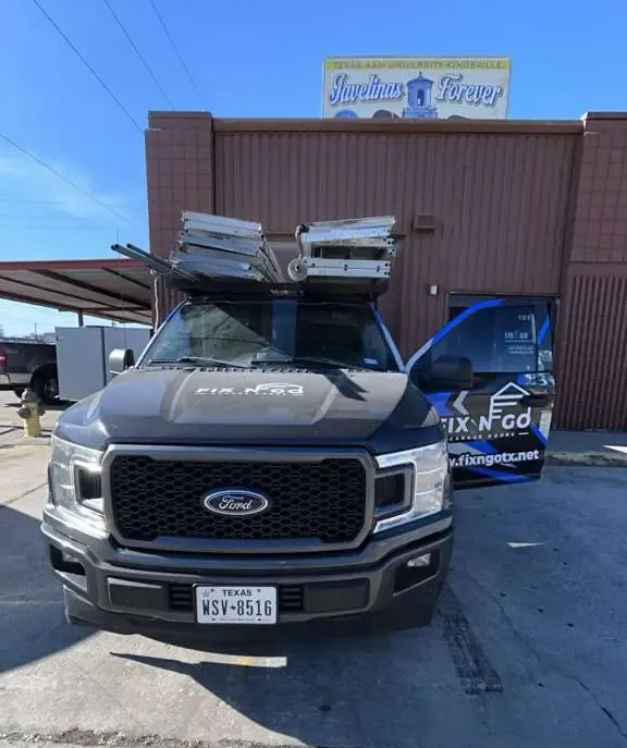 Black Ford pickup with luggage on top parked outside a brick building and blue van.