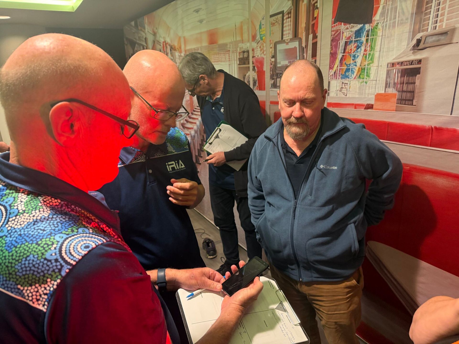 Four men examining an object indoors. One man shines a red light, others hold papers and a phone.