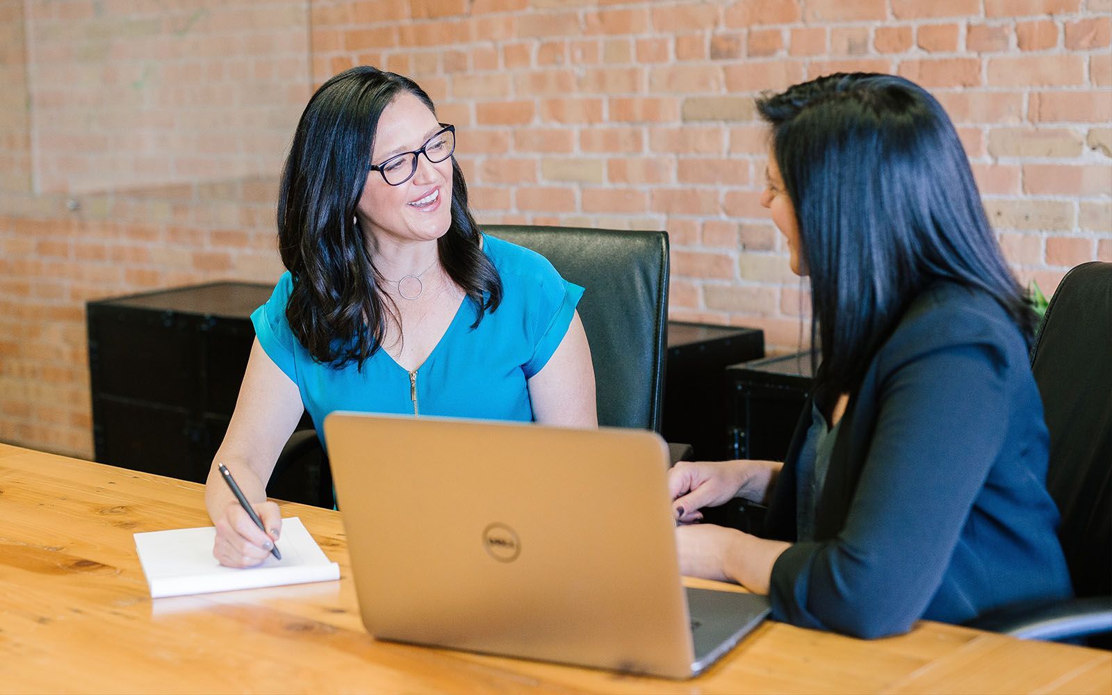 Two women in discussion at a table; one writes on a pad, the other looks at a laptop.