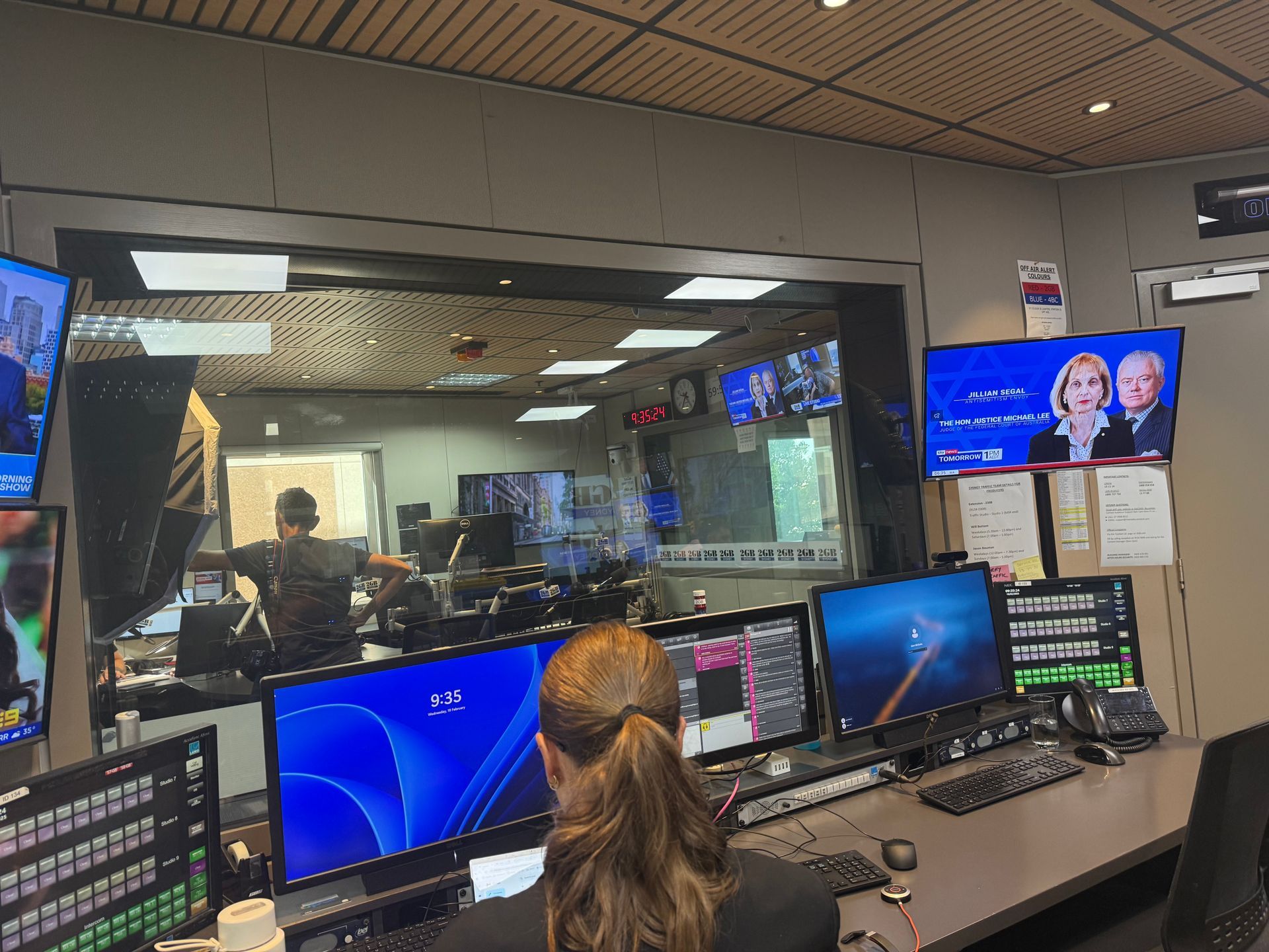 Inside a broadcast control room; a person works at a desk in the foreground. Monitors and people in the background.