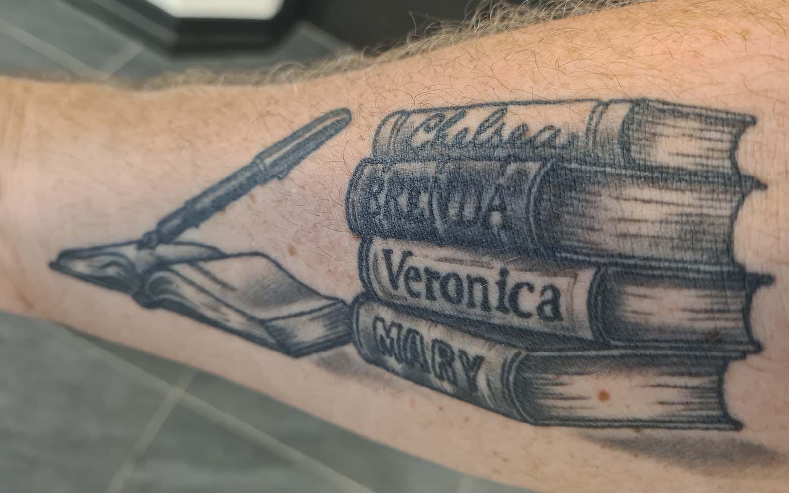 Tattoo of four books with names on the spines: Chelsea, Silvia, Veronica, and Mary, plus a pen and book.