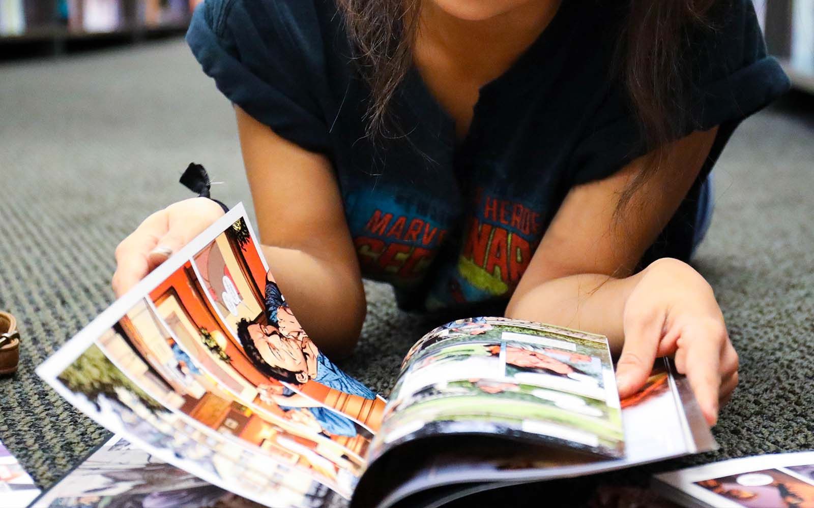 Person reading a comic book on the floor of a library, surrounded by bookshelves.