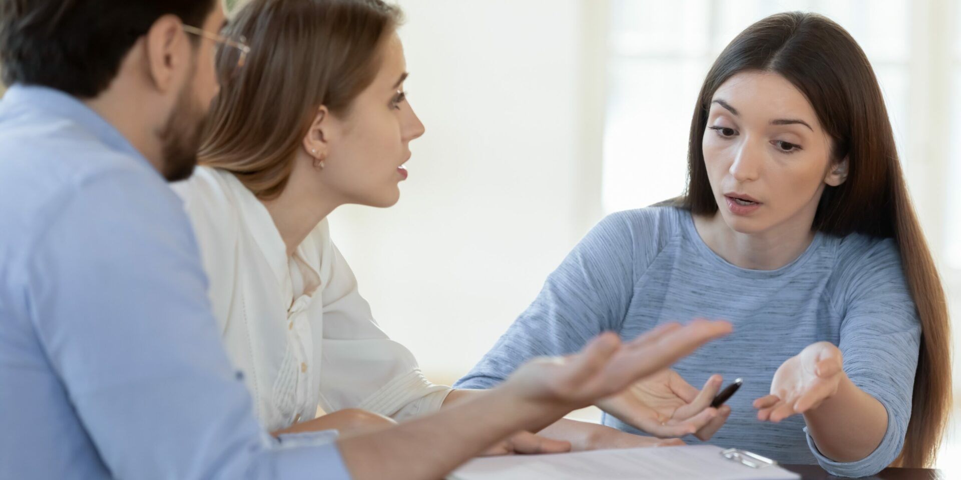 Three people in discussion, one gesturing, looking at the others. They are in a well-lit room.