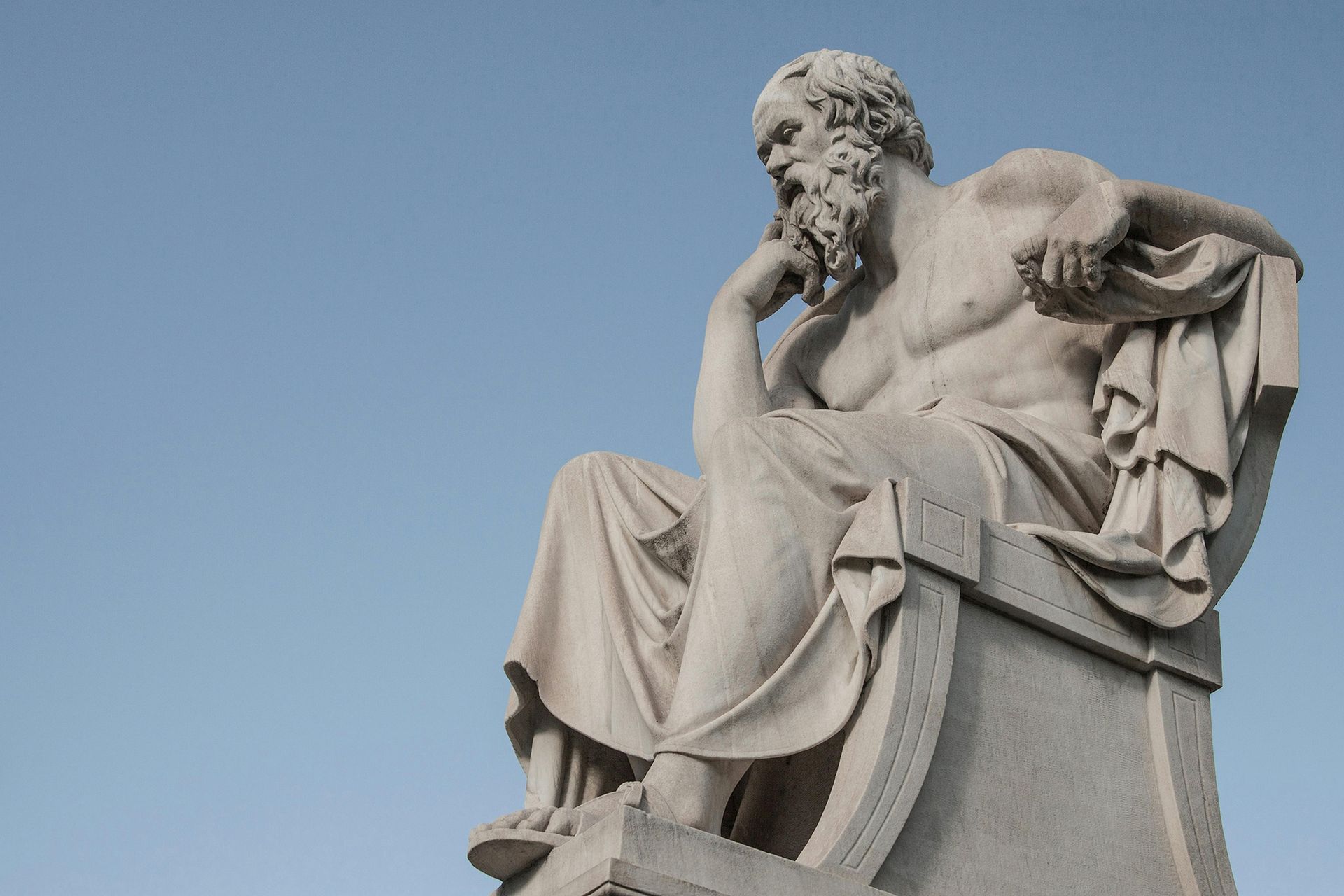 Statue of a seated, thoughtful philosopher with a beard, against a blue sky.