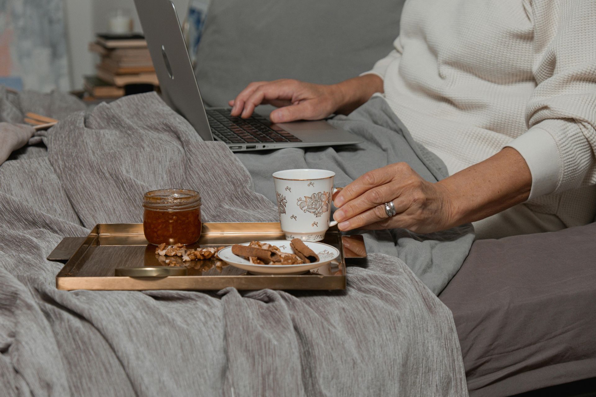Person using a laptop in bed, holding a mug, with a tray of treats nearby.