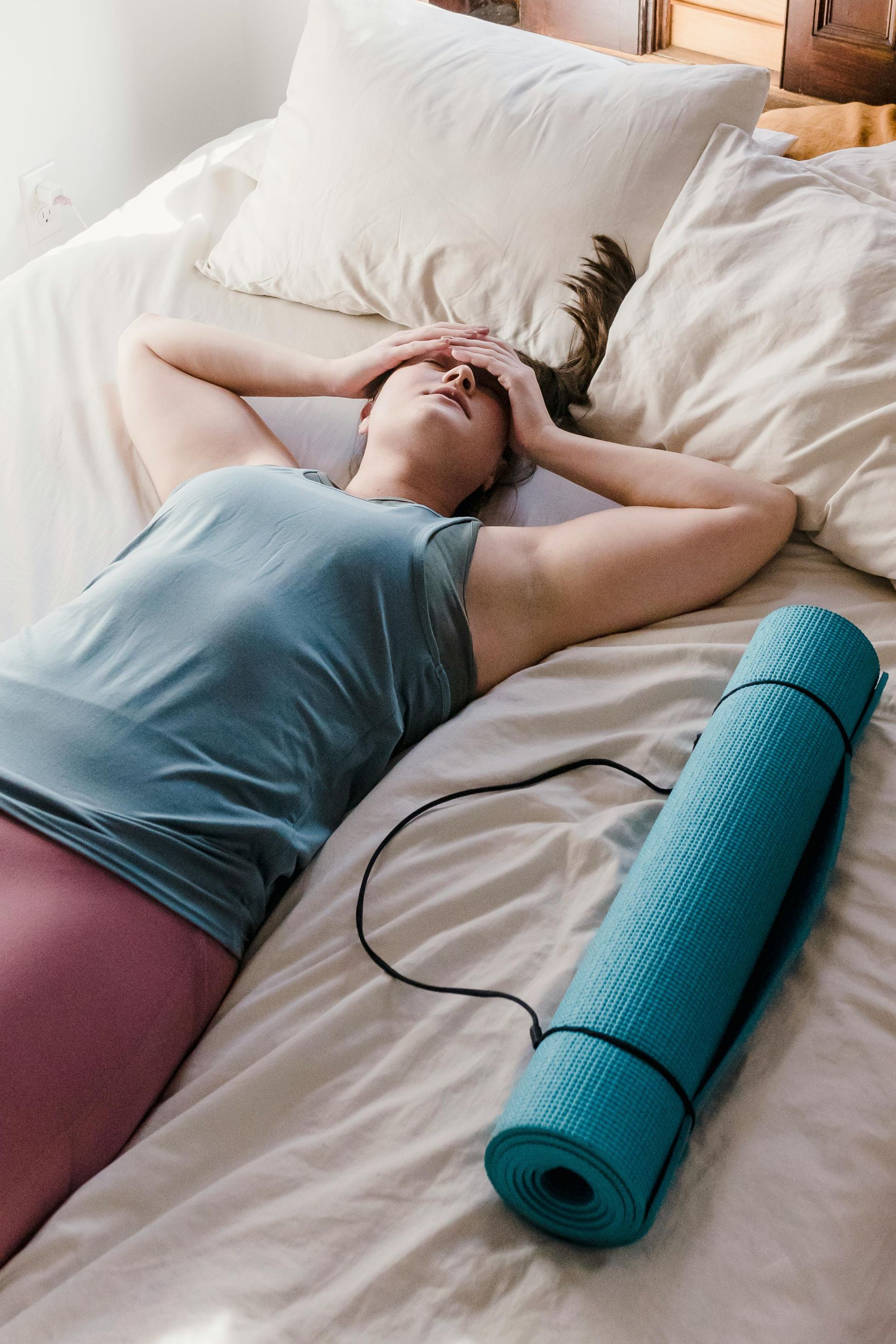 Woman in workout clothes lying on bed, appearing tired. Yoga mat nearby.