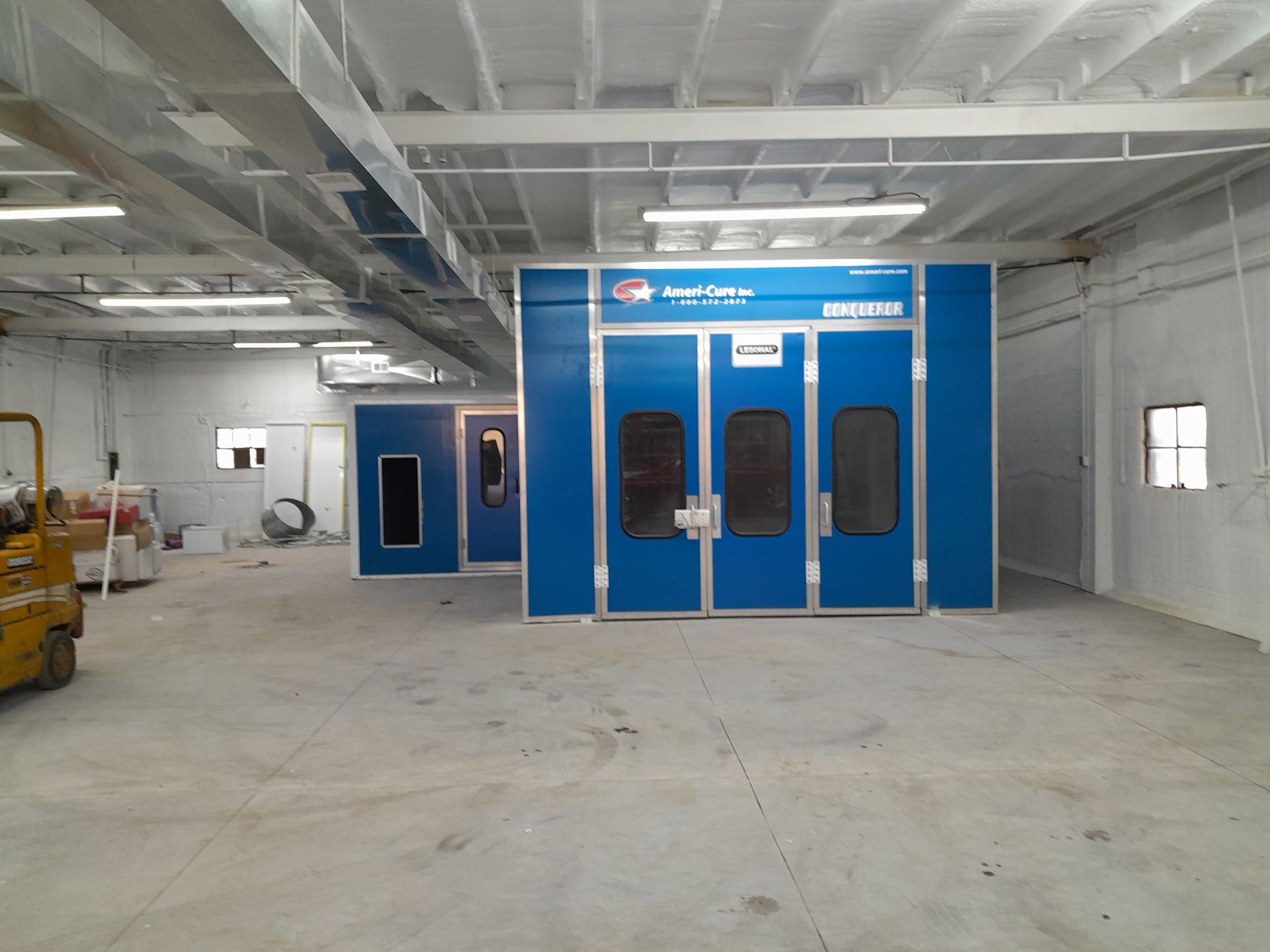 A blue paint booth in a warehouse with a yellow forklift in the background