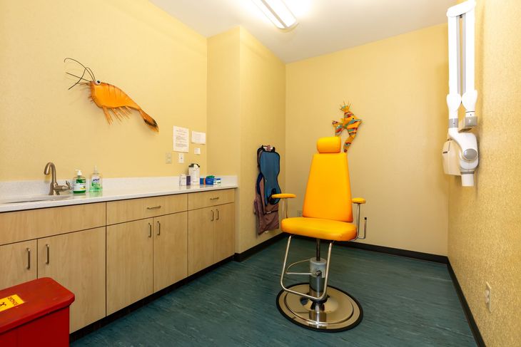 Empty dental exam room with yellow chair, cabinets, sink, and wall-mounted equipment