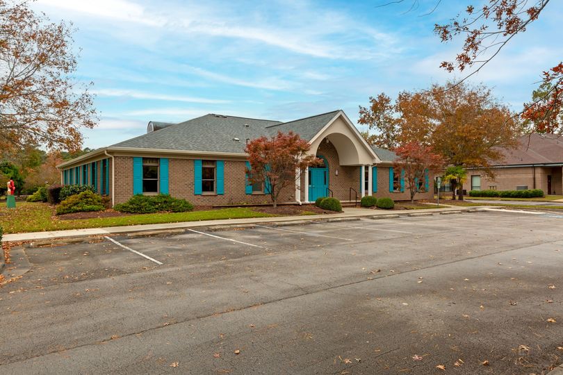 Brick office building with teal doors and windows, fronted by a mostly empty parking lot and autumn trees