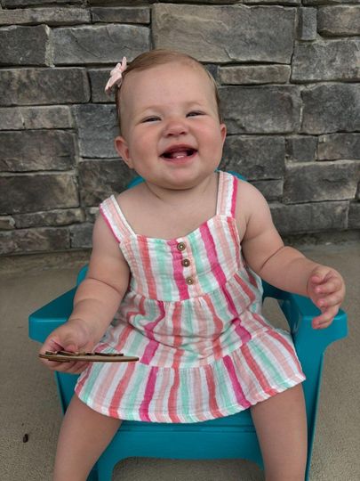 Smiling baby in a striped romper sitting on a blue chair against a stone wall.