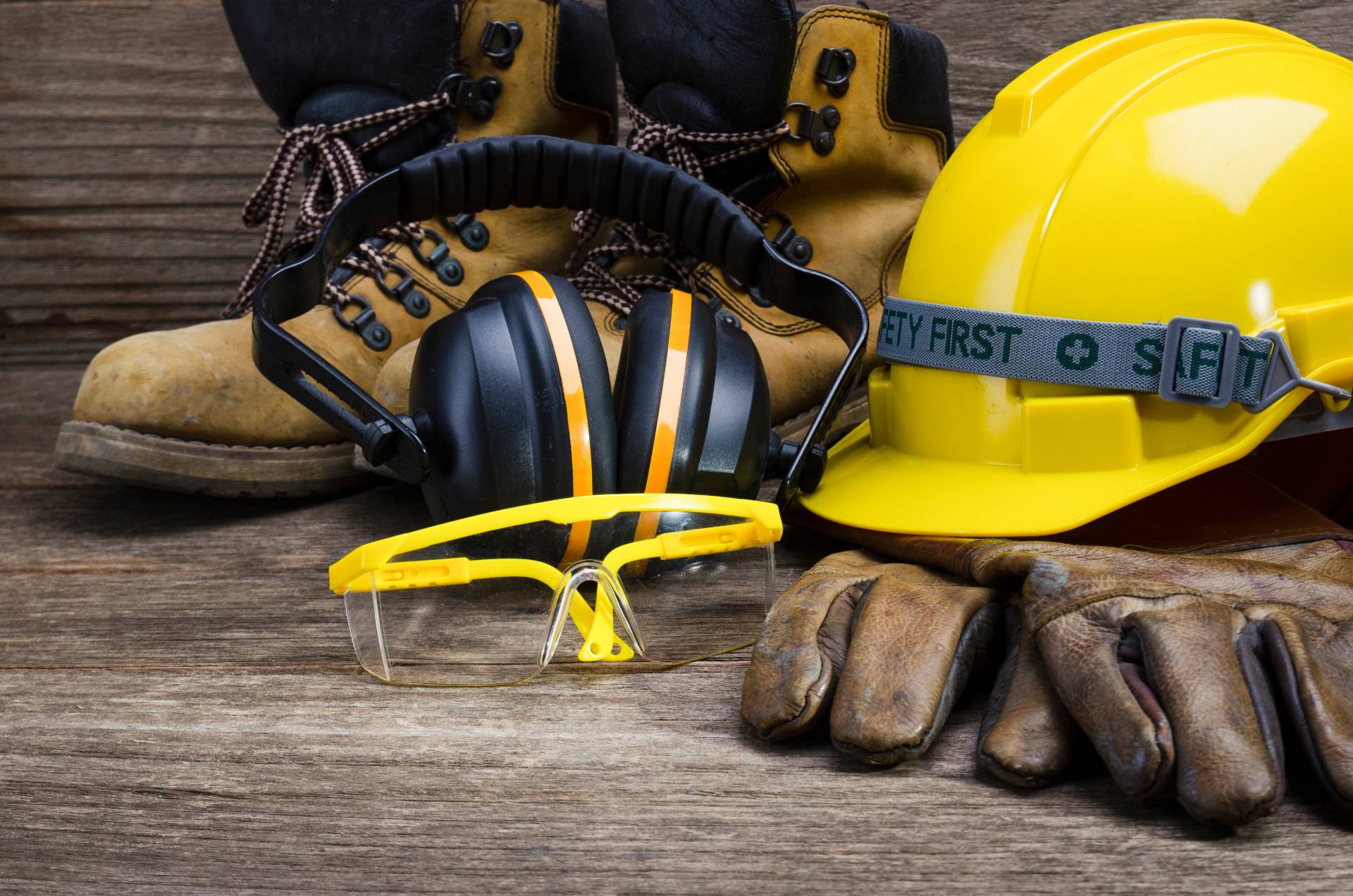 Su un tavolo di legno sono presenti un elmetto protettivo, cuffie antirumore, occhiali di sicurezza, guanti e stivali.