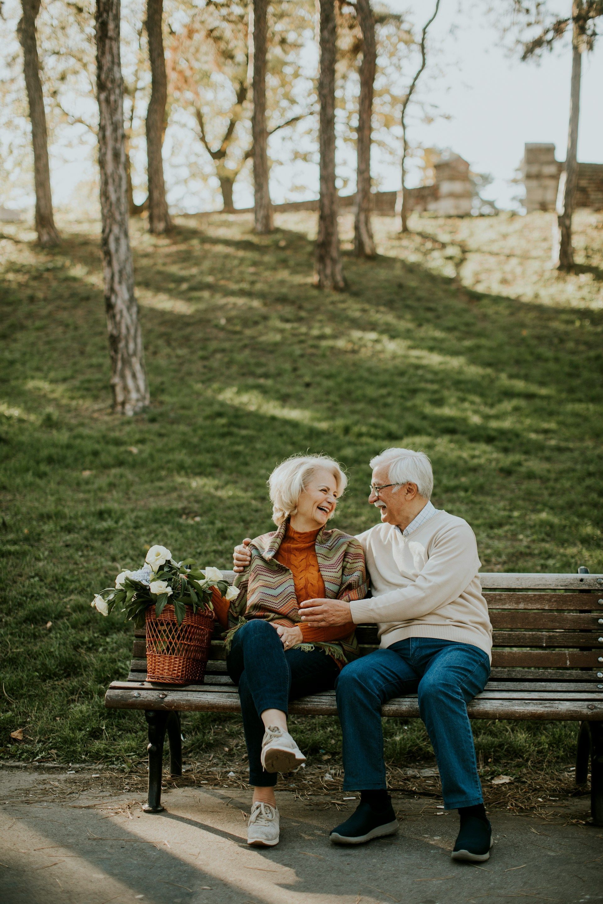 Elderly couple laughing and embracing on park bench with basket of flowers.