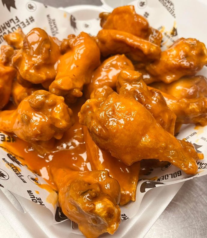 A pile of chicken wings heavily coated in orange buffalo sauce, served in a food tray lined with branded paper.