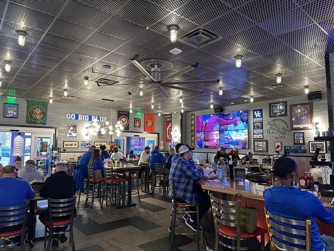 Patrons sit at tables and a bar in a sports bar with a metallic, patterned ceiling, large TV, and blue decor.