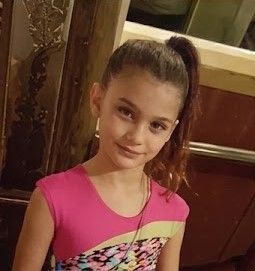 Girl in a pink dress smiling indoors, standing by wooden cabinets.