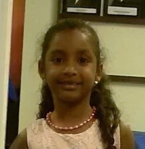 Smiling child in a pink dress and necklace, standing indoors against a wall and cabinet.