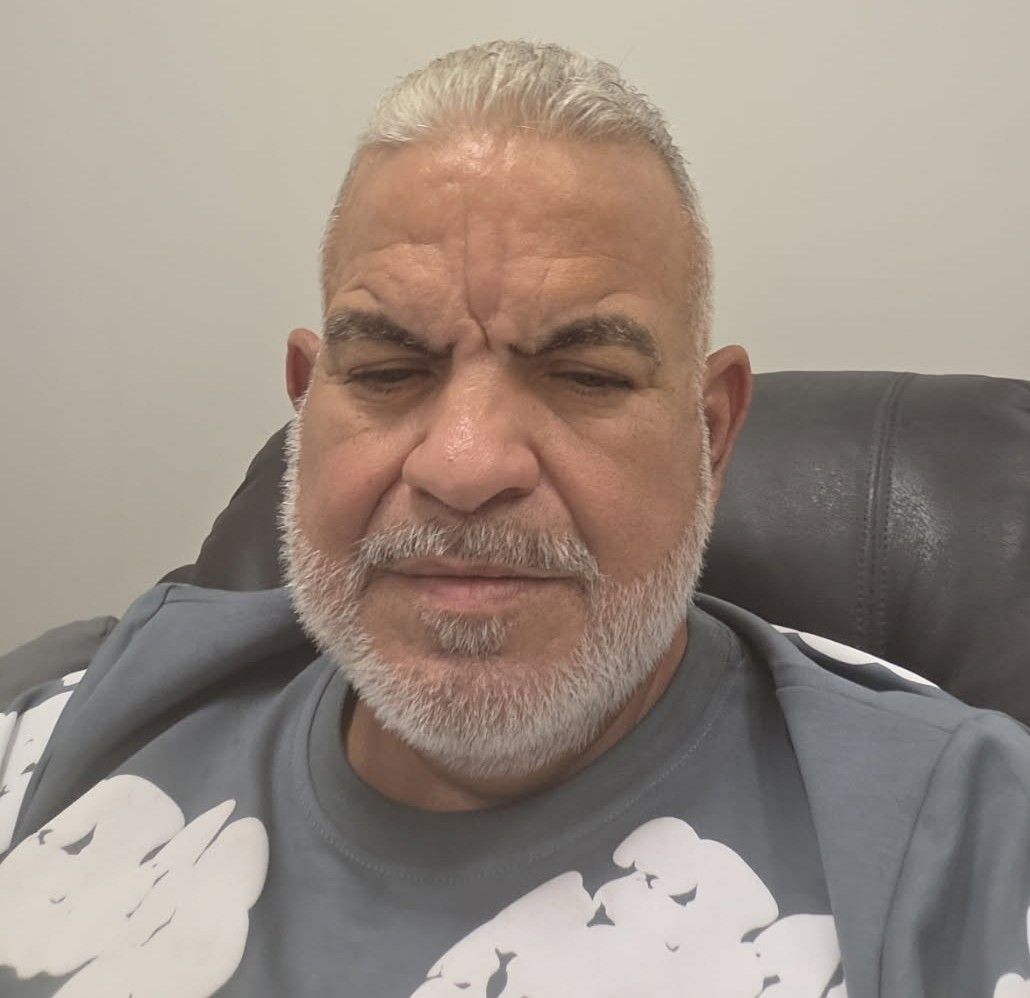 Man with gray hair and beard, wearing a gray shirt, seated on a dark leather chair with eyes closed.