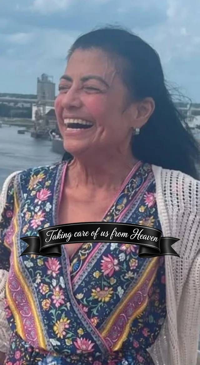 Woman laughing outdoors by the water, wearing a colorful embroidered top and white shawl.
