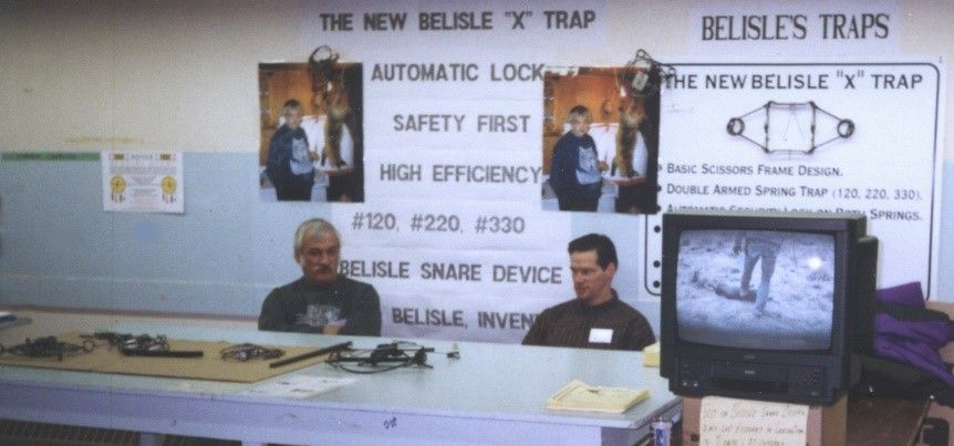 Deux hommes sont assis à une table, des affiches publicitaires pour « Belisle's Traps » et des composants de pièges.