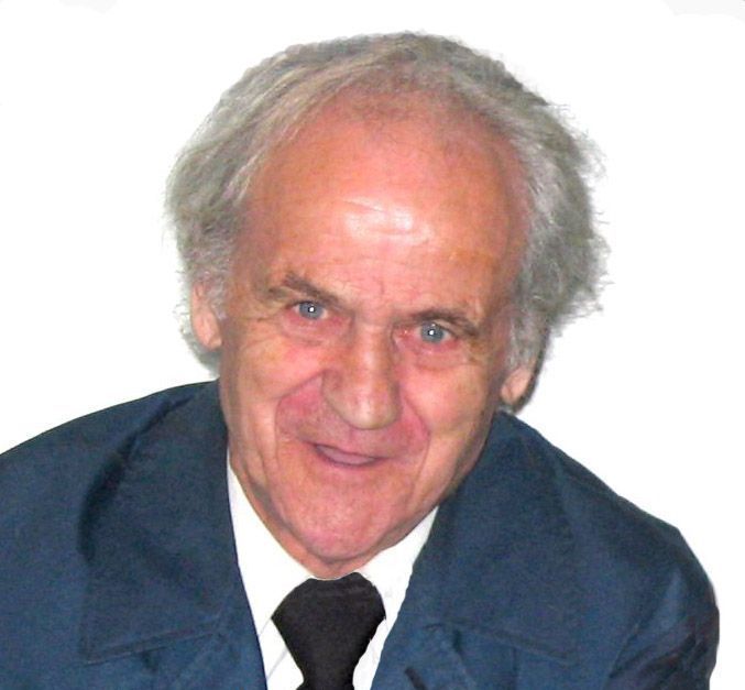 Un homme âgé souriant, aux cheveux gris, portant une veste sombre et une cravate noire.