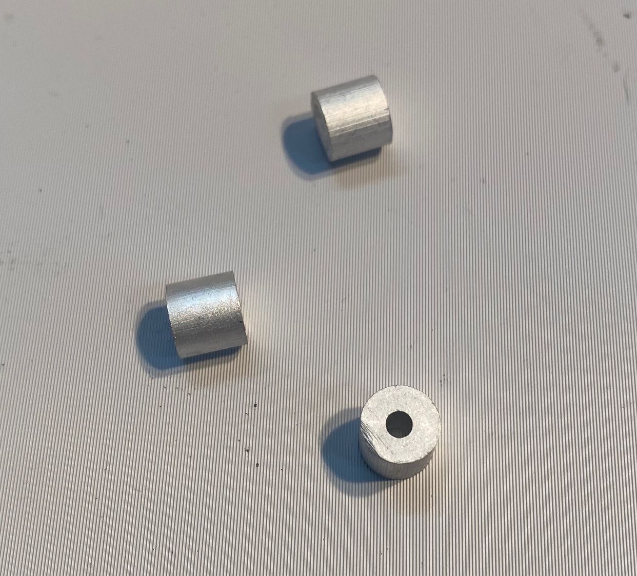 Trois petites entretoises cylindriques argentées sur une surface grise texturée. L'une d'elles présente un trou central.