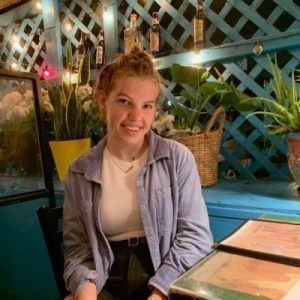 A smiling person wearing a light blue button-up shirt sits at a table in a dimly lit restaurant with a blue lattice wall.