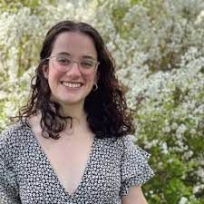 A smiling person with curly brown hair and glasses wearing a patterned top in front of blooming white trees.